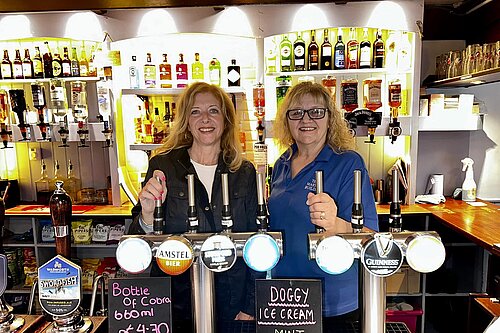 Liz Jarvis MP behind the bar with a smiling staff member at a local pub. Both women are standing in front of beer taps, with shelves of spirits behind them.