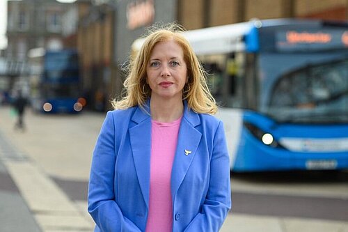 Liz Jarvis, MP for Eastleigh, stands in a blue jacket in front of a bus, she has a neutral expression. 